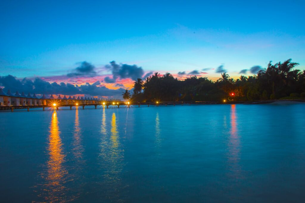 Serene sunset view of water villas and palm trees at Hakuraa Huraa, Maldives, reflecting vibrant colors on the sea.