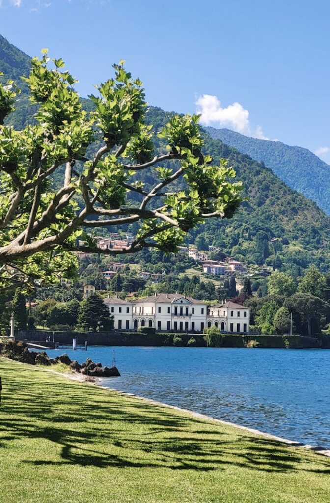 Beautiful view of a villa by the lake, framed by trees and mountains in a summer setting.